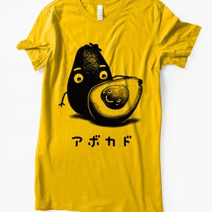 Peut inclure: Un t-shirt jaune avec une illustration en noir et blanc de deux avocats avec des visages. Le texte "アボカド" est imprim&eacute; sous les avocats.