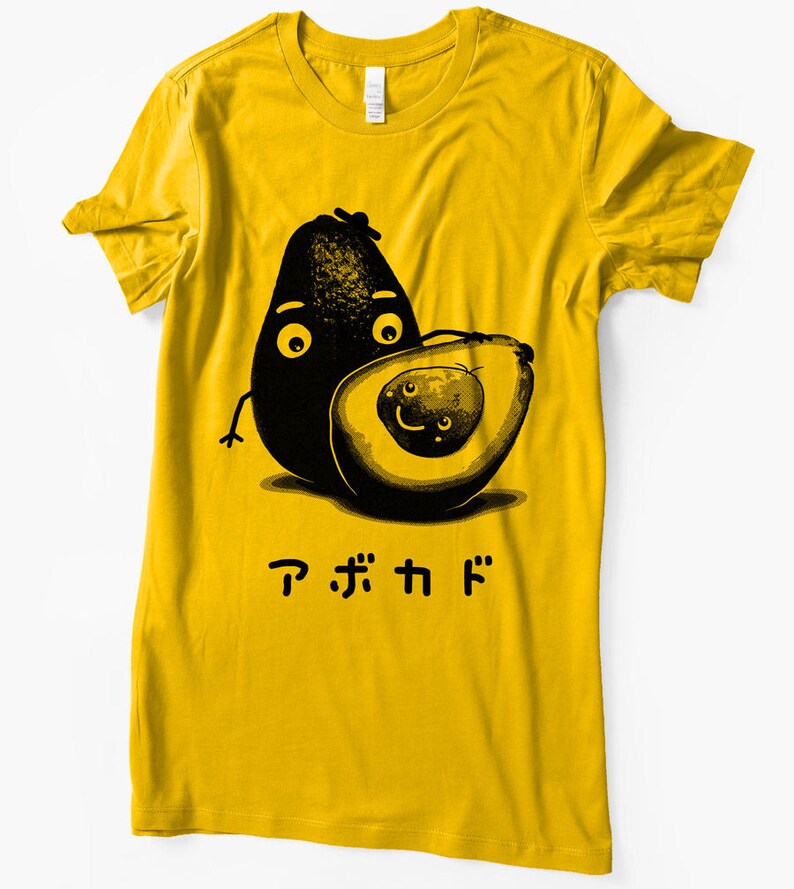 Peut inclure: Un t-shirt jaune avec une illustration en noir et blanc de deux avocats avec des visages. Le texte "アボカド" est imprim&eacute; sous les avocats.
