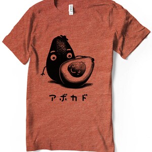Peut inclure: Un t-shirt rouge avec un motif d'avocat dessin&eacute;. L'avocat a un visage et tient un plus petit avocat. Le texte "アボカド" est imprim&eacute; sous les avocats.