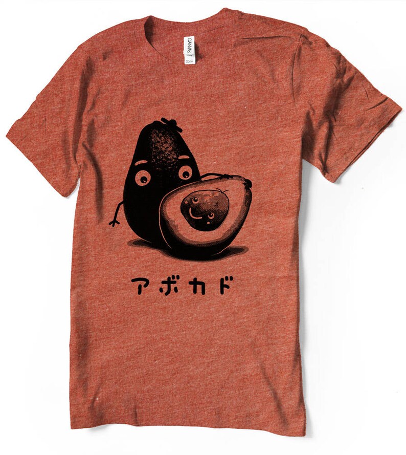 Peut inclure: Un t-shirt rouge avec un motif d'avocat dessin&eacute;. L'avocat a un visage et tient un plus petit avocat. Le texte "アボカド" est imprim&eacute; sous les avocats.