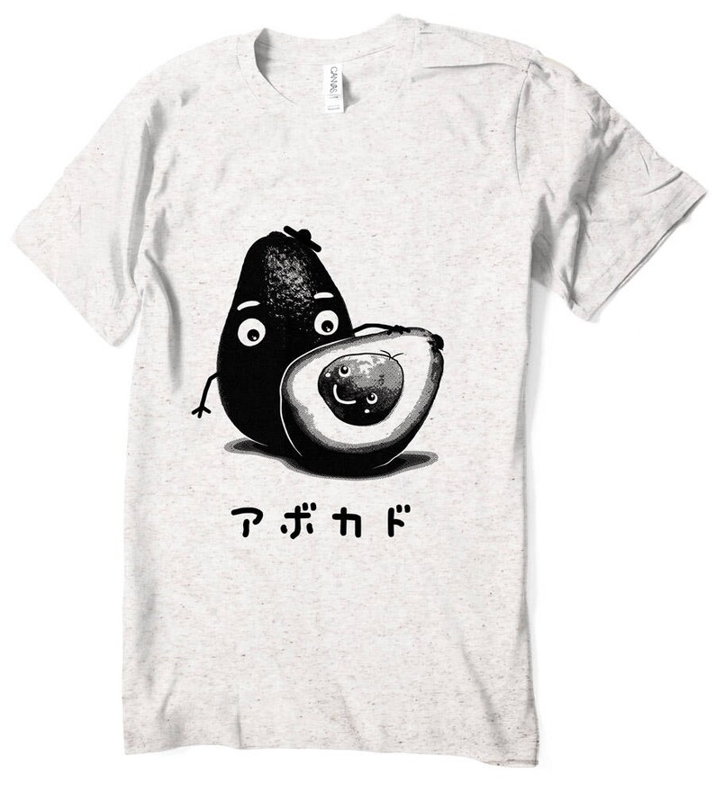 Peut inclure: Un t-shirt blanc avec une illustration en noir et blanc de deux avocats avec des visages. L'avocat de gauche est entier et l'avocat de droite est coup&eacute; en deux. Le texte "アボカド" est imprim&eacute; sous les avocats.