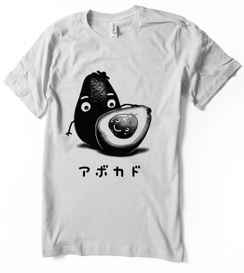 Peut inclure: Un t-shirt blanc avec une illustration en noir et blanc de deux avocats avec des visages. L'avocat &agrave; gauche a une expression surprise et l'avocat &agrave; droite sourit. Le texte "アボカド" est imprim&eacute; sous les avocats.