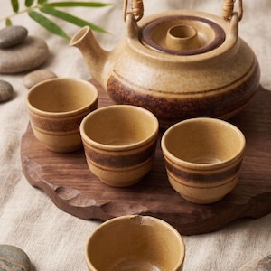 May include: A ceramic tea set featuring a teapot with a bamboo handle and four matching cups. The pieces are light tan with dark brown horizontal bands. The teapot has a lid with a central knob, and one cup shows a small chip on the rim.