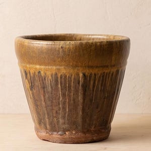 May include: A ceramic planter with a wide, rounded rim and a tapered body. The exterior features a two-tone glaze with a golden-brown upper section that transitions into a darker, mottled brown base with vertical drip patterns and ridges.