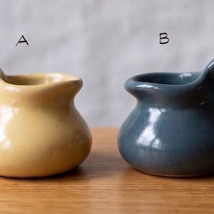 May include: Two small ceramic pitchers with rounded bodies and single side handles. The pitcher labeled A is a solid light yellow color, while the pitcher labeled B is a solid slate blue color. Both items feature a smooth, glossy finish.