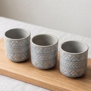 May include: Three cylindrical ceramic cups featuring a light gray base color and a repeating geometric pattern of horizontal bands with blue and beige chevron and arrow motifs. The cups have a smooth interior finish and a textured exterior surface.