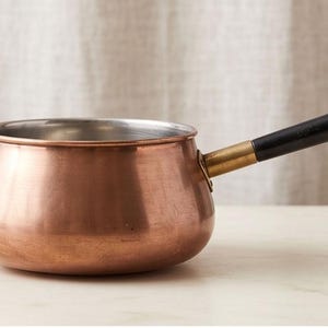 May include: A copper saucepan with a rounded body and a silver-toned interior. The pot features a long, straight black handle attached to the side with a brass-colored metal ferrule, extending outward from the main body of the cooking vessel.