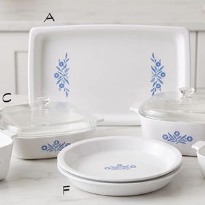 May include: A collection of white ceramic bakeware featuring a blue floral pattern. The set includes a rectangular platter labeled A, a square dish B, a lidded rectangular dish C, a lidded rectangular dish D, a small square dish E, and round dishes F.