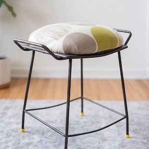 May include: A stool with a dark metal frame and four thin legs featuring small yellow caps at the base. The seat is round and padded, upholstered in a fabric with a pattern of large, overlapping circles in shades of cream, gray, and olive green.