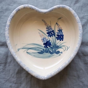 May include: A heart-shaped ceramic bowl with a cream-colored interior featuring a hand-painted blue floral motif of grape hyacinth flowers. The rim of the bowl has a speckled light blue sponge-painted pattern against a cream background.