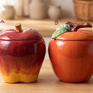 May include: Two ceramic apple-shaped canisters with lids. Canister A is deep red fading to yellow at the base with a brown stem. Canister B is orange with a green leaf and a brown stem. Both feature glossy finishes and are labeled A and B.