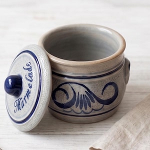 May include: A grey ceramic jar with a matching lid featuring a dark blue hand-painted scroll design and the word Marmelade written in blue script. The jar has a smooth interior, a textured lid surface, and a dark blue knob handle on the lid.
