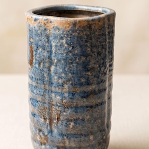 May include: A cylindrical ceramic vase with a textured, mottled blue glaze featuring a crackled finish. The surface displays vertical ridges and exposed brown clay patches along the rim and sides, creating a weathered appearance with a wide opening.