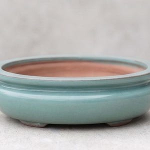 May include: A round, shallow ceramic bonsai pot with a smooth, glossy teal-green glaze on the exterior and a matte, unglazed terracotta-colored interior. The vessel features a thick, rounded rim and rests on small, integrated feet at the base.