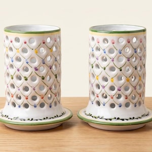 May include: Two cylindrical white ceramic candle holders featuring a lattice cut-out pattern. The exterior is decorated with a hand-painted green, yellow, and pink grid design, with a green leaf motif encircling the base and a yellow rim.