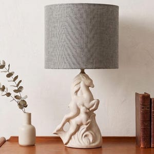 May include: A table lamp featuring a cream-colored ceramic base sculpted into the shape of a rearing horse and a smaller foal. The lamp is topped with a cylindrical lampshade covered in a textured, grey fabric with a subtle herringbone pattern.
