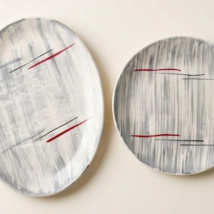 May include: An oval plate labeled A and a round plate labeled B, both featuring a white base with vertical grey brushstroke patterns and horizontal red and black accent lines. The plates are arranged side by side on a plain light background.