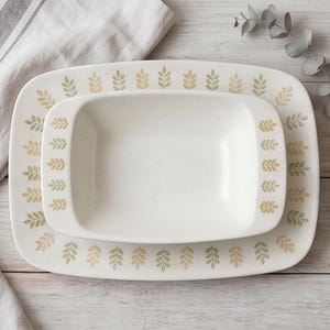 May include: A rectangular ceramic serving platter and a matching rectangular serving bowl, both featuring an off-white base with a speckled finish and a repeating pattern of small, alternating sage green and tan leaf sprigs along the edges.