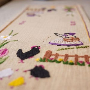 May include: A rectangular beige fabric table runner featuring colorful embroidery of a person in a purple bonnet carrying a basket, a brown wooden fence, a black chicken, a white daisy, a pink tulip, and small yellow and white birds.