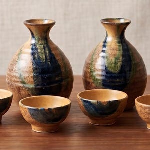 May include: A set of two ceramic sake bottles and four matching small cups. Each piece features a textured, earthy tan base with a prominent, vertical dark blue and green glaze drip pattern running down the center of the rounded vessels.