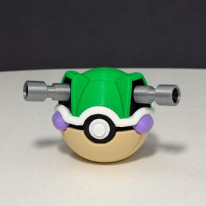May include: A spherical toy featuring a green top half and a tan bottom half, separated by a white band with a central black and white circular button. Two silver cylindrical cannons extend from the sides, and two small purple accents are visible.