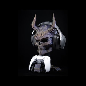 May include: A skull-shaped headphone stand with horns, a headset, and a white game controller. The skull is metallic with purple and bronze tones. The headset is black and gray. The controller is white with black buttons.