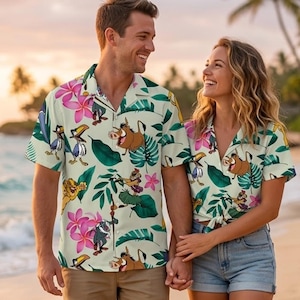 Puede incluir: Camisas con botones de manga corta a juego con un estampado tropical con personajes de dibujos animados, flores rosas y hojas verdes. Las camisas las lleva una pareja en la playa. Las camisas son de color verde claro.