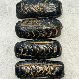 May include: Four cylindrical black beads arranged vertically. Each bead features a textured surface with a repeating carved herringbone or chevron pattern running lengthwise, revealing lighter brown material within the recessed grooves.