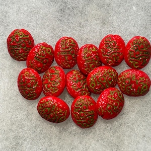 May include: A collection of thirteen oval-shaped beads, each featuring a vibrant red base color with an intricate, raised green pattern of swirling lines and small dots covering the surface of every individual bead in the set.