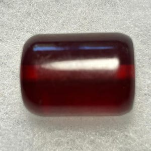 May include: A smooth, cylindrical bead with a deep, translucent dark red color. The surface is polished and reflective, showing a horizontal band of lighter red light passing through the center of the bead, highlighting its semi-transparent nature.
