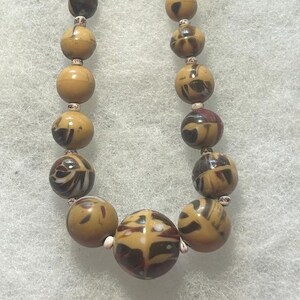 May include: A necklace featuring two strands of spherical beads in shades of tan, dark brown, and cream. The beads exhibit a marbled, swirled pattern with a glossy finish. Small, speckled off-white spacer beads are positioned between the larger beads.
