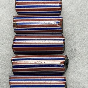 Puede incluir: Cinco cuentas de vidrio vintage con forma cilíndrica. Cada cuenta presenta rayas alternas rojas, blancas y azules. Las cuentas están dispuestas verticalmente sobre un fondo claro, resaltando su diseño a rayas. Miden aproximadamente 2 cm de largo.
