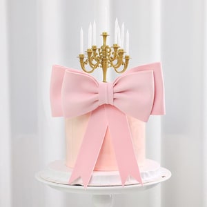 May include: A pink cake with a large pink bow and a gold candelabra cake topper with white candles. The cake sits on a white cake stand against a white backdrop.