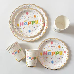 以下が含まれることがあります： 金色のスカラップエッジが付いた、お祝い用の食器セット。白いお皿とカップには、カラフルな文字で「Happy」の文字、風船、旗、紙吹雪が描かれています。