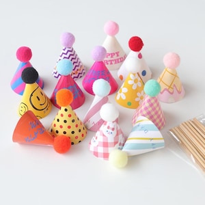 May include: A collection of colorful party hats with pom-poms. The hats feature various patterns, including stripes, polka dots, and floral designs. Some hats have text that reads "Happy Birthday" and "Happy Day".