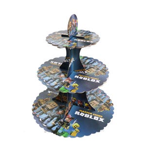 Puede incluir: Un soporte para cupcakes de cart&oacute;n de tres niveles con tem&aacute;tica de Roblox. El soporte tiene una combinaci&oacute;n de colores azul y blanco con personajes de Roblox y las palabras "Master Skins Roblox" impresas en cada nivel. Los bordes de los niveles son festoneados.