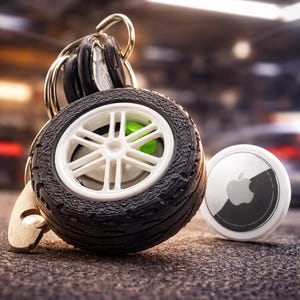 Könnte beinhalten: Ein Schlüsselanhänger mit einem Miniatur-Reifen in Schwarz, einer weißen Felge und einem leuchtend grünen Bremssattel, befestigt an einem Metallring mit Schlüsseln. Daneben liegt ein weißer Apple AirTag mit silbernem Logo auf der Vorderseite.
