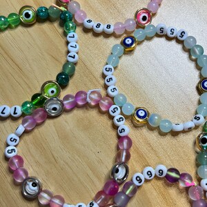 May include: Several beaded bracelets in shades of pink, green, and blue. Each bracelet features a combination of round beads, white letter beads, and decorative eye charms. The bracelets are arranged on a light brown wooden surface.
