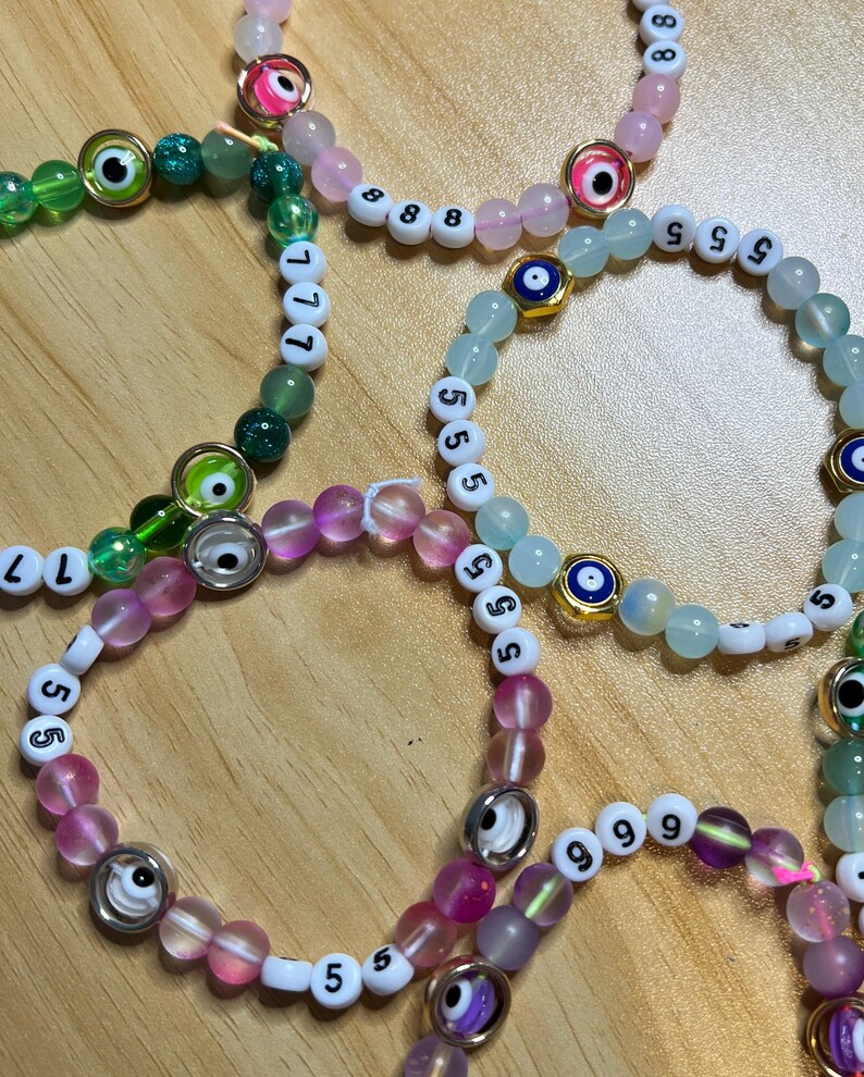 May include: Several beaded bracelets in shades of pink, green, and blue. Each bracelet features a combination of round beads, white letter beads, and decorative eye charms. The bracelets are arranged on a light brown wooden surface.