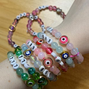 May include: A collection of beaded bracelets in various colors, including pink, green, and white. Some bracelets feature numbers and evil eye charms. The beads are made of glass and other materials. The bracelets are worn on a wrist.