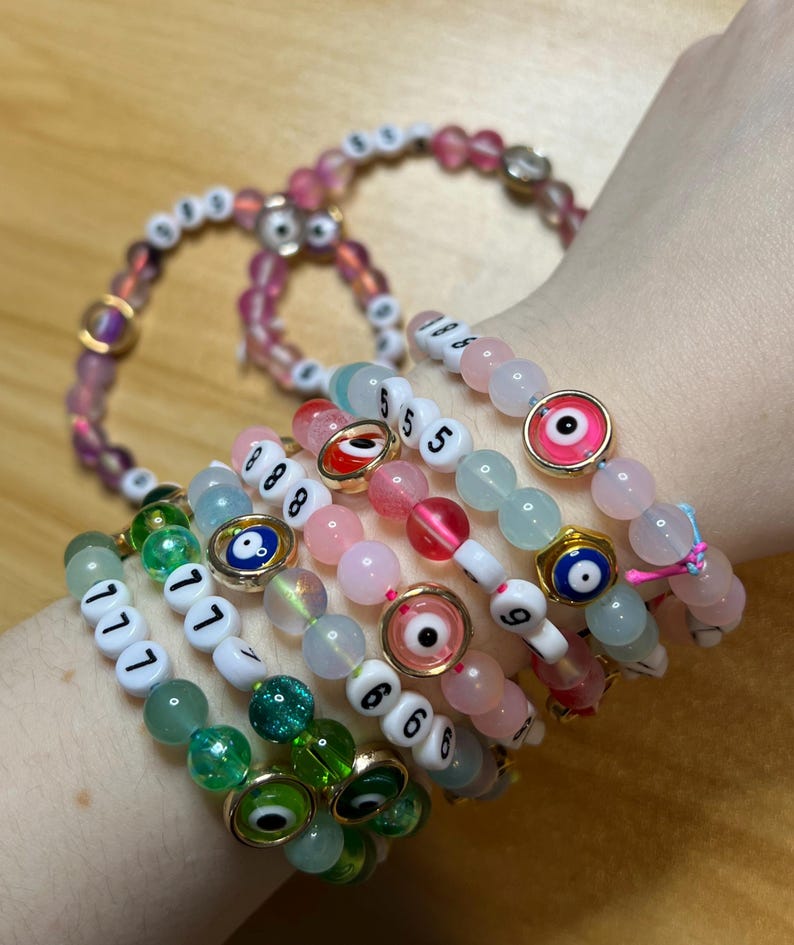 May include: A collection of beaded bracelets in various colors, including pink, green, and white. Some bracelets feature numbers and evil eye charms. The beads are made of glass and other materials. The bracelets are worn on a wrist.