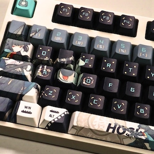 May include: A custom mechanical keyboard with a cream-colored frame and a variety of keycap designs. The keycaps feature anime-style artwork, geometric patterns, and text. The keyboard has a mix of dark gray, light blue, and white keys.