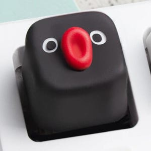 May include: A black keycap with a cartoon penguin face design. The keycap features white eyes and a red beak. The keycap is on a white keyboard, with other white keycaps visible in the background.
