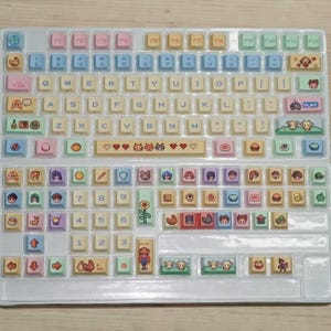 May include: A custom keyboard with pastel-colored keycaps. The keycaps feature various icons and illustrations, including hearts, animals, and food items. The keyboard layout is standard, with function keys and a number pad. The keycaps are arranged in a clear plastic tray.