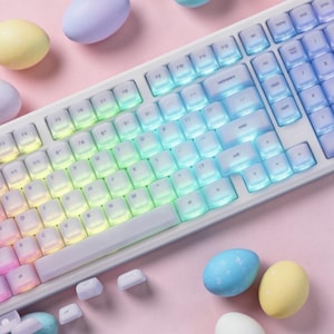 May include: A white mechanical keyboard with rainbow-colored backlit keys. The keys are arranged in a standard layout. Pastel-colored Easter eggs are scattered around the keyboard on a pink surface. The keyboard has a modern, minimalist design.
