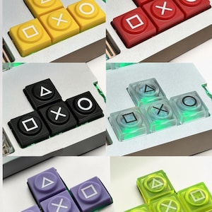 Könnte beinhalten: Eine Sammlung von individuell gestalteten Gaming-Tastenkappen in verschiedenen Farben, darunter Gelb, Rot, Schwarz, Transparent, Lila und Grün. Jedes Set zeigt die ikonischen Dreieck-, Kreis-, X- und Quadrat-Symbole, die man von Gamecontrollern kennt. Die Tastenkappen sind auf einer silbernen Basis angeordnet.