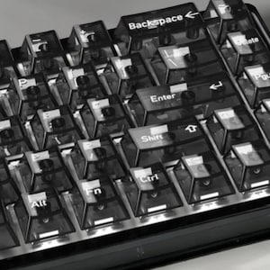 Może przedstawiać: Zbliżenie klawiatury mechanicznej z przezroczystymi nakładkami na klawisze. Klawisze są ułożone w standardowym układzie QWERTY, z widocznymi etykietami takimi jak "Backspace", "Enter", "Shift" i "Ctrl". Klawiatura ma nowoczesny design.
