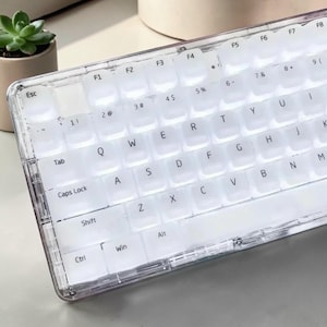 Może przedstawiać: Przezroczysta klawiatura z białymi klawiszami. Klawiatura ma standardowy układ z literami, cyframi i klawiszami funkcyjnymi. Klawisze są podświetlane, a klawiatura ma przezroczystą ramkę.