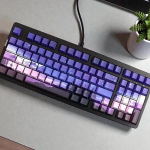 Op de afbeelding: Een zwart mechanisch toetsenbord met aangepaste keycaps met een gradiëntontwerp van paars, blauw en wit, dat lijkt op een landschap met een sterrenhemel. Het toetsenbord heeft een bedrade verbinding.