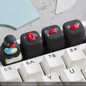May include: A set of custom black penguin-themed keycaps with red beaks and white eyes, some with tears. One keycap features a penguin holding a blue fish. The keycaps are on a white keyboard.